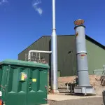 Tall steel chimney connected to a large green warehouse with generator